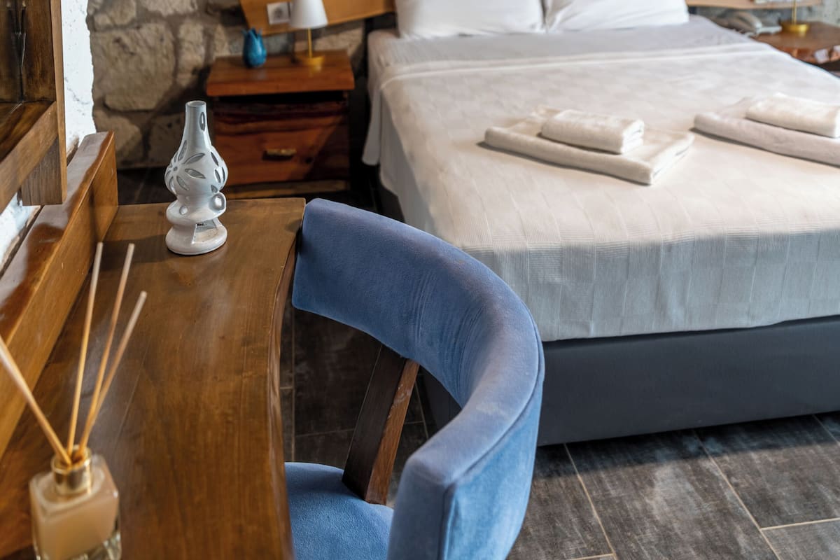 A bedroom with a stone wall, wooden headboard, a bed with white linens, a nightstand with a lamp, and a blue chair.