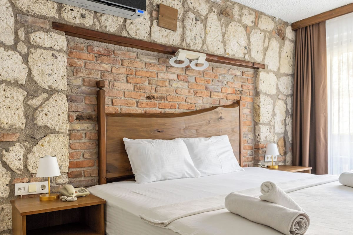 A hotel room with a brick wall, a wooden headboard, a bed with white linens, a nightstand with a lamp, and a wall-mounted air conditioner.