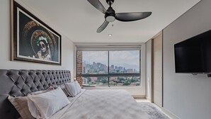 Une chambre avec vue sur la ville, un ventilateur de plafond, un lit recouvert d’une courtepointe à motifs et une œuvre d’art encadrée accrochée au mur.