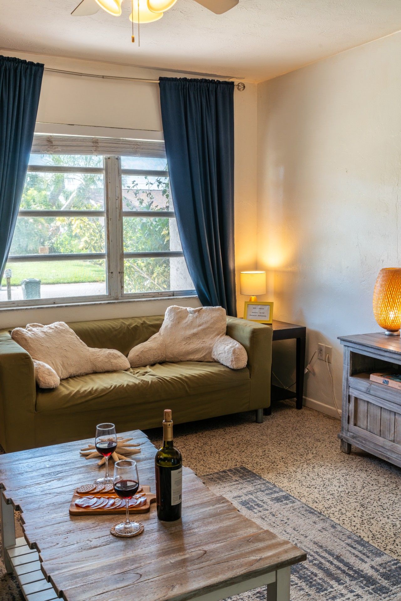 A cozy living room with a sofa, a coffee table with wine glasses and a bottle, a lamp, and a view of greenery outside.