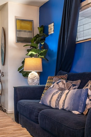 A room with a blue wall, a dark blue sofa with patterned cushions, a round table with a lamp, and a framed artwork. - Private Coastal Haven: Fire Pit • Yard • Beach (Sarasota)