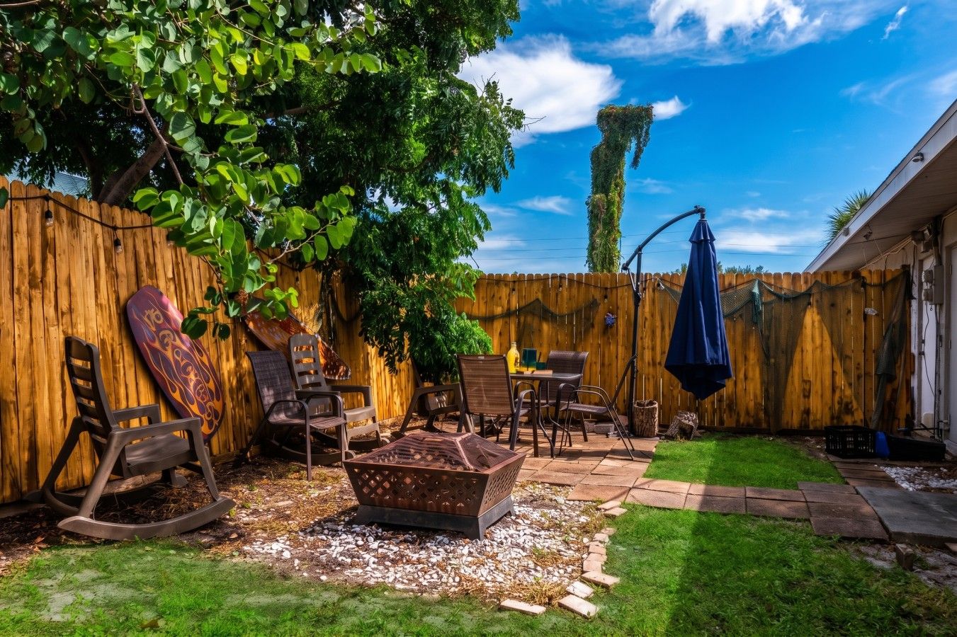 A backyard with a wooden fence, a patio table with chairs, a decorative metal planter, and a blue umbrella.