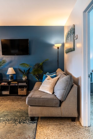 A living room with a sofa, a flat-screen TV mounted on the wall, a floor lamp, and a painting. - Chic Local Hideaway: BBQ Nights • Near Beach (Sarasota)