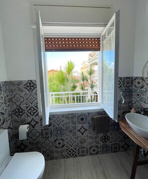 Un baño con baldosas estampadas, inodoro, lavamanos y ventana con vista al balcón.