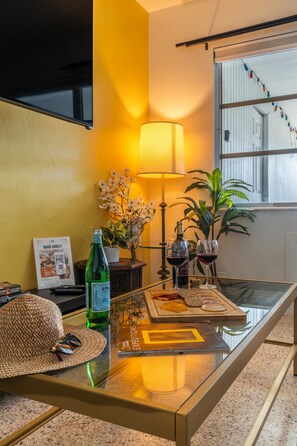 Ein Zimmer mit einem Glastisch, einer Lampe, einer Flasche, einem Weinglas, einem Hut, einem Buch und einer Pflanze.