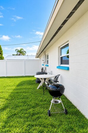 A backyard with a grill, a table, and a white fence. - Chic Sarasota Gem: Beach • BBQ • Workspace • Patio (Sarasota)