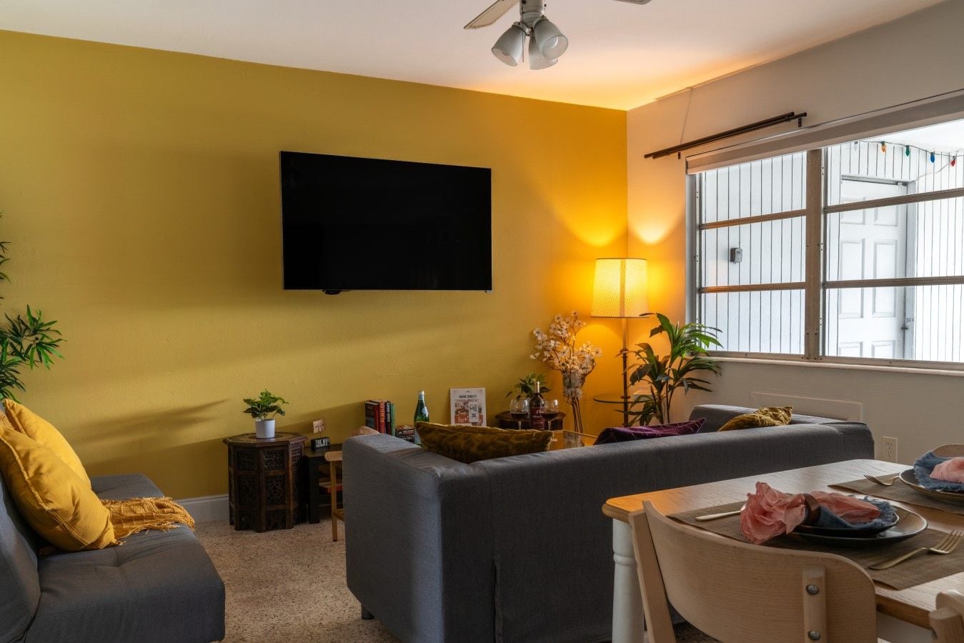 A living room with a sofa, a dining table set, a TV mounted on the wall, and a window with blinds.