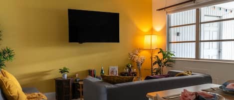 Una sala de estar con sofá, mesa de comedor, televisor montado en la pared y ventana con persianas.