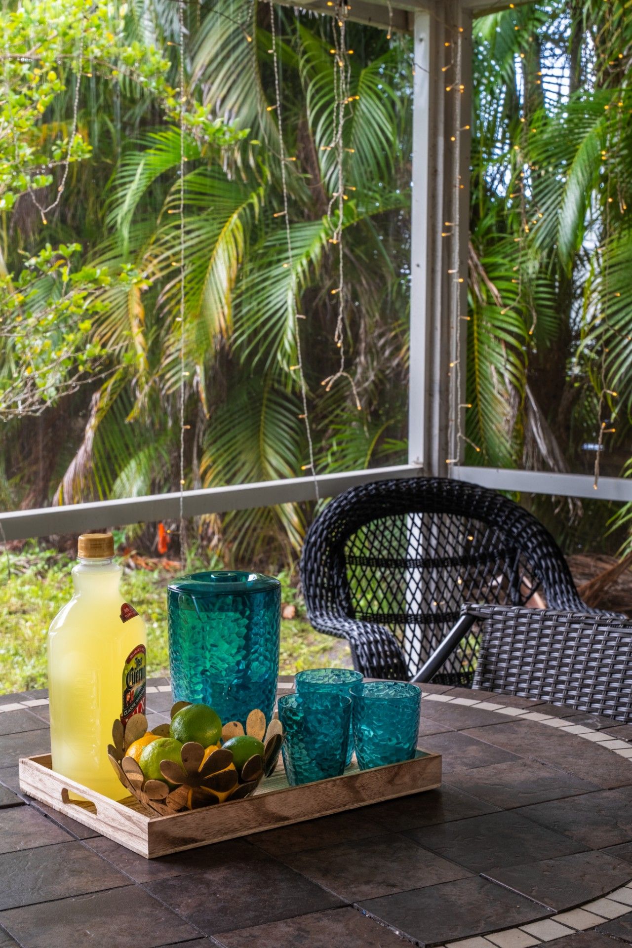 A tropical outdoor seating area with a wicker chair, a tray with a bottle and glasses, and lush greenery in the background.