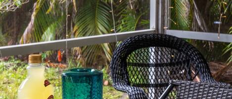 A tropical outdoor seating area with a wicker chair, a tray with a bottle and glasses, and lush greenery in the background.