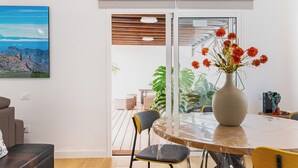 Un comedor con una mesa redonda, sillas, un florero con flores y vistas a un espacio exterior con plantas a través de un ventanal corredero.