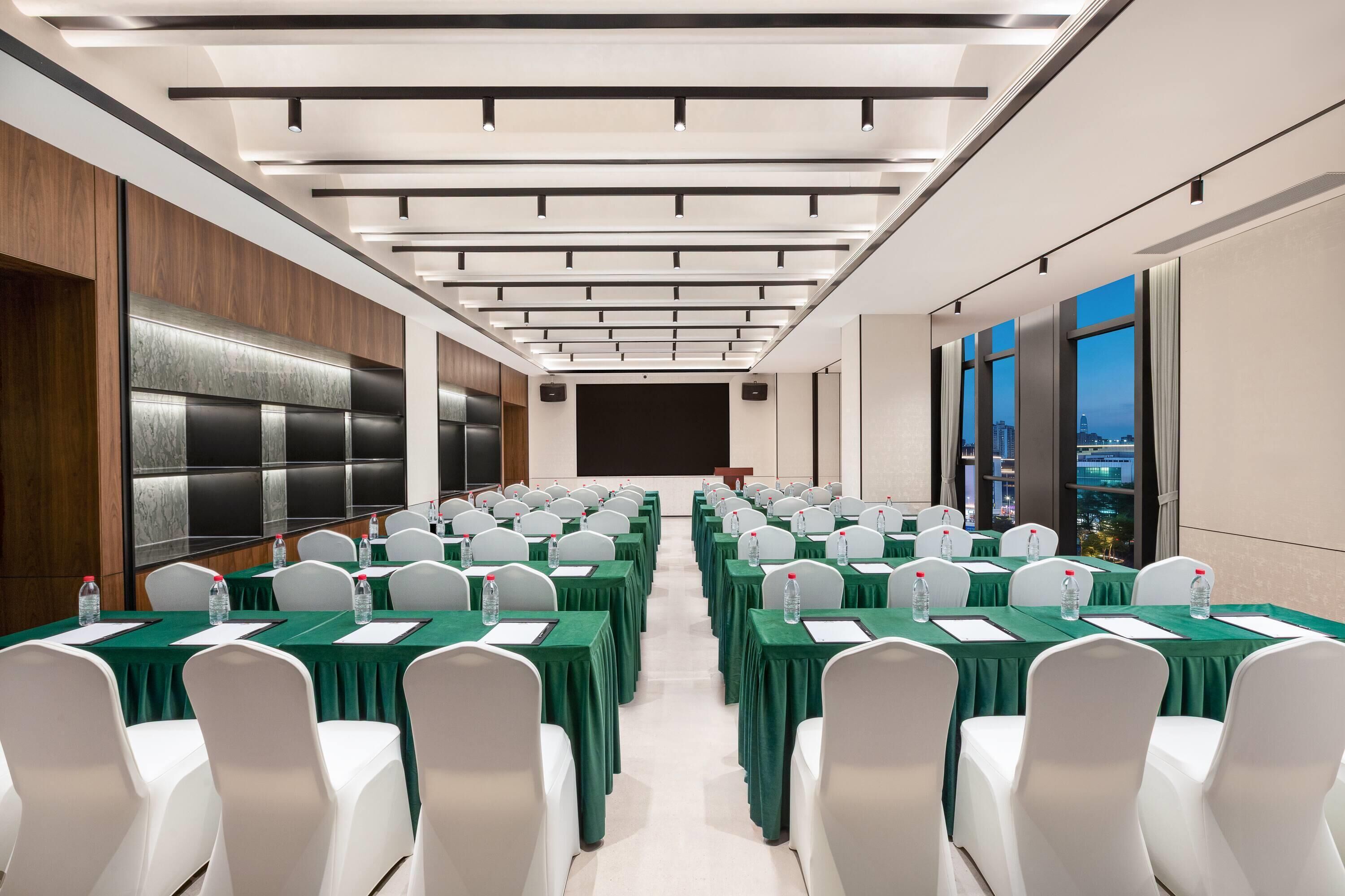 A conference room with rows of tables, white chairs, and a large screen at the front.
