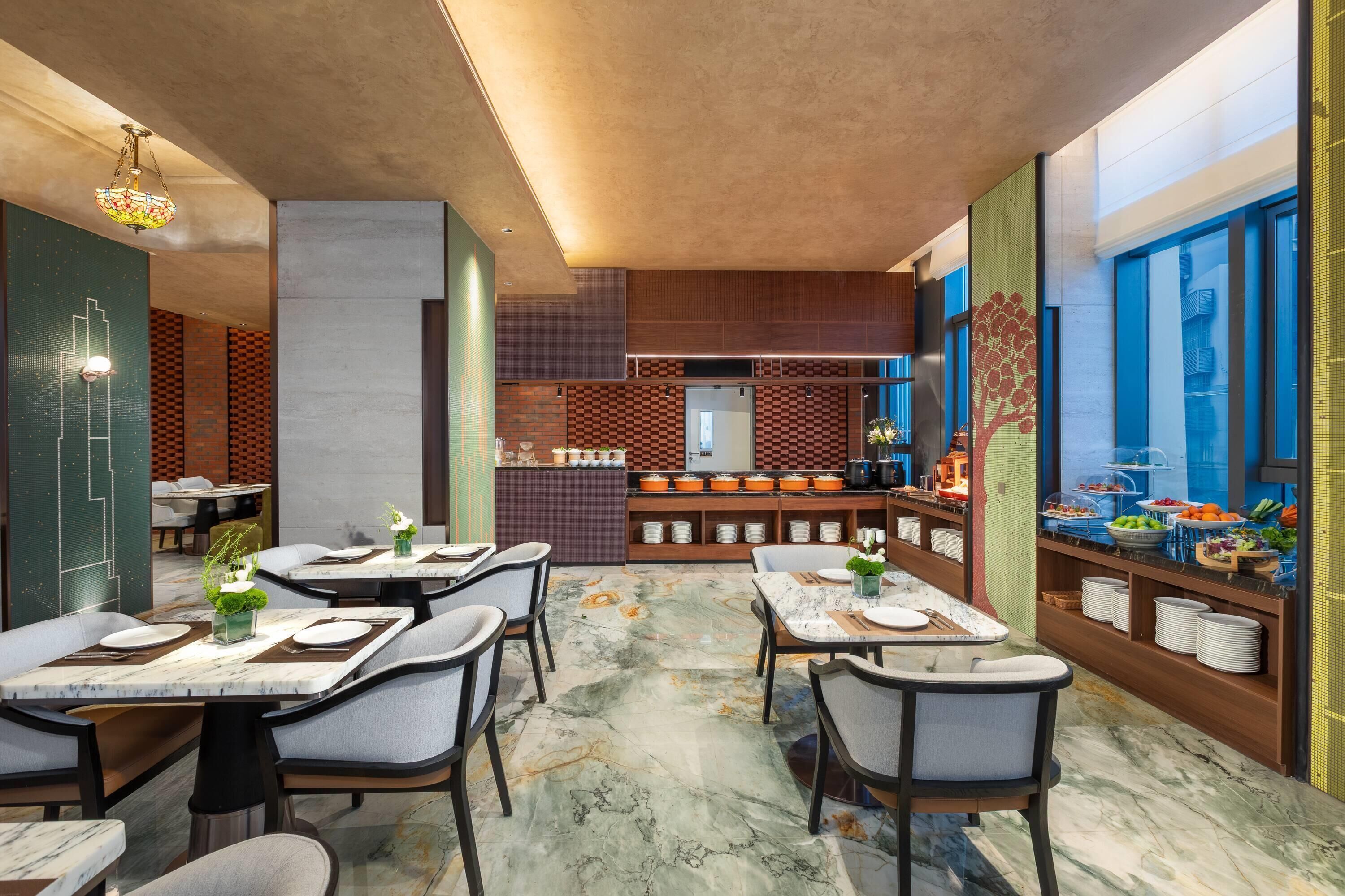 A modern restaurant with marble flooring, grey upholstered chairs, and a buffet area with various food items.