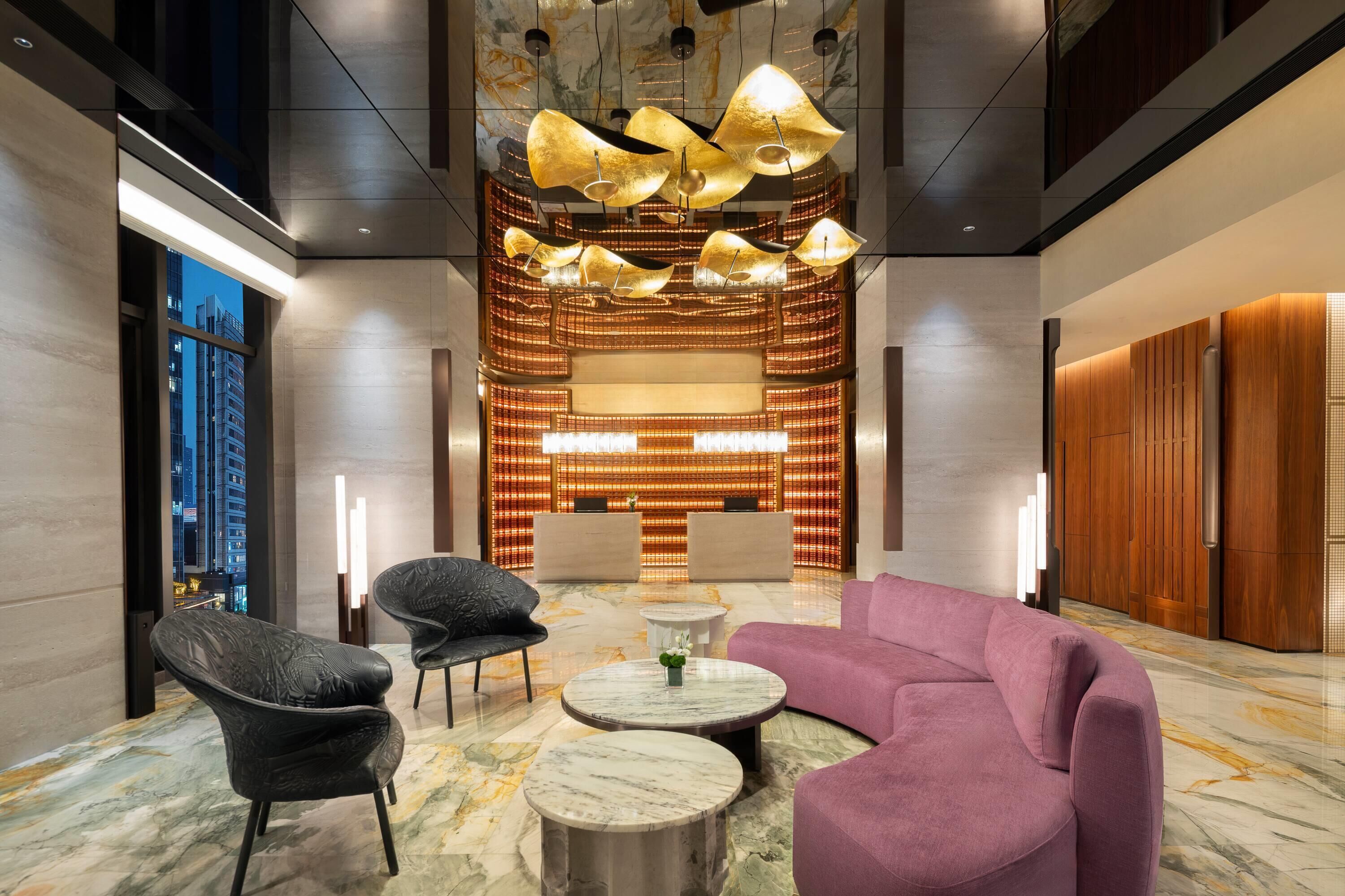 A lobby with a marble floor, a purple sofa, black chairs, a central table, and a decorative light fixture.