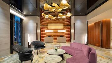 A lobby with a marble floor, a purple sofa, black chairs, a central table, and a decorative light fixture.