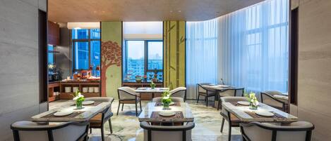 A modern dining area with multiple tables and chairs, large windows with city views, and a mix of natural and artificial lighting.