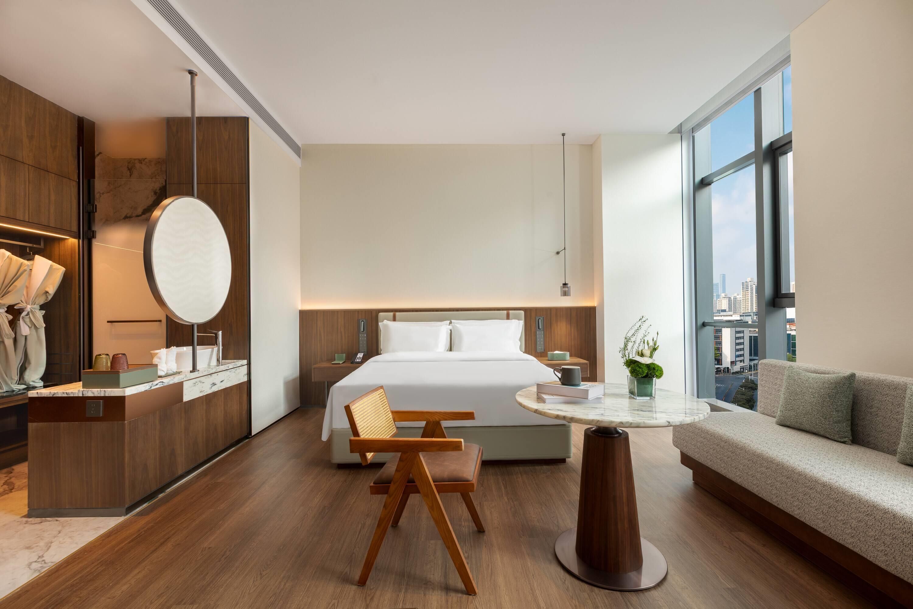 Habitación de hotel moderna con cama, sofá, mesa de comedor, silla, espejo y vistas a la ciudad.