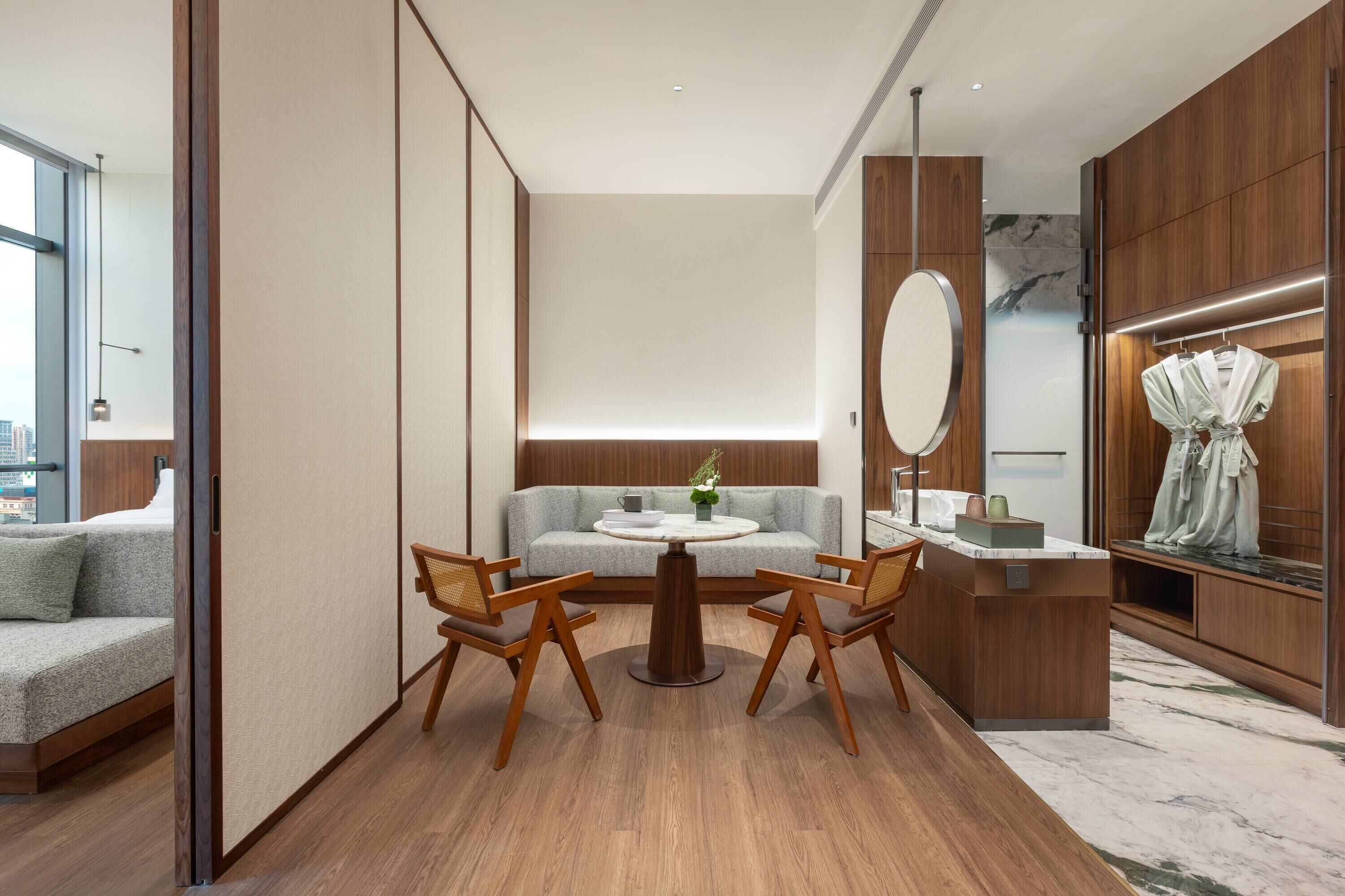 A modern hotel room with a sofa, a dining table with chairs, a small kitchen area, and a large mirror.