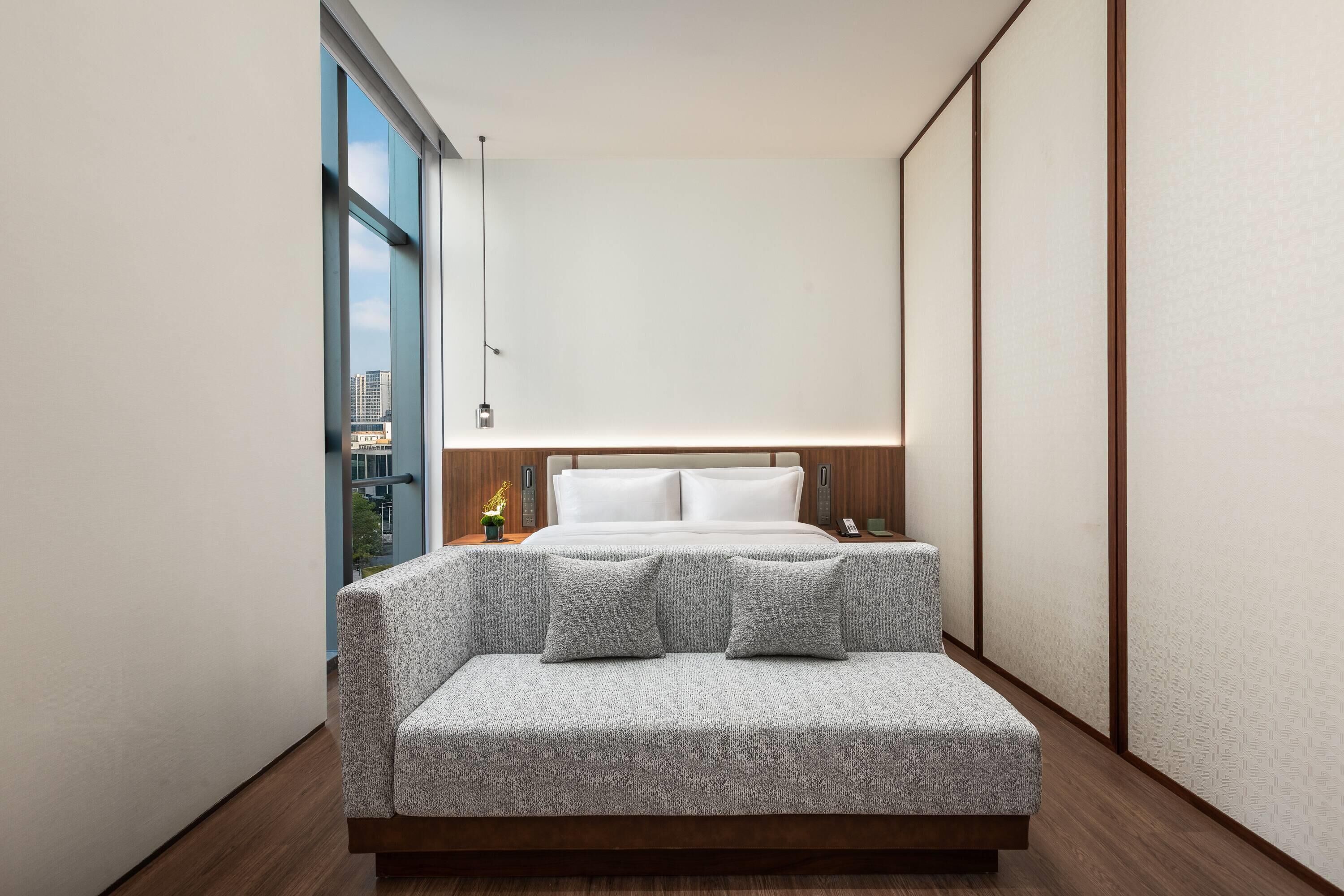 A modern hotel room with a bed, a sofa, a nightstand, and a view of the city.