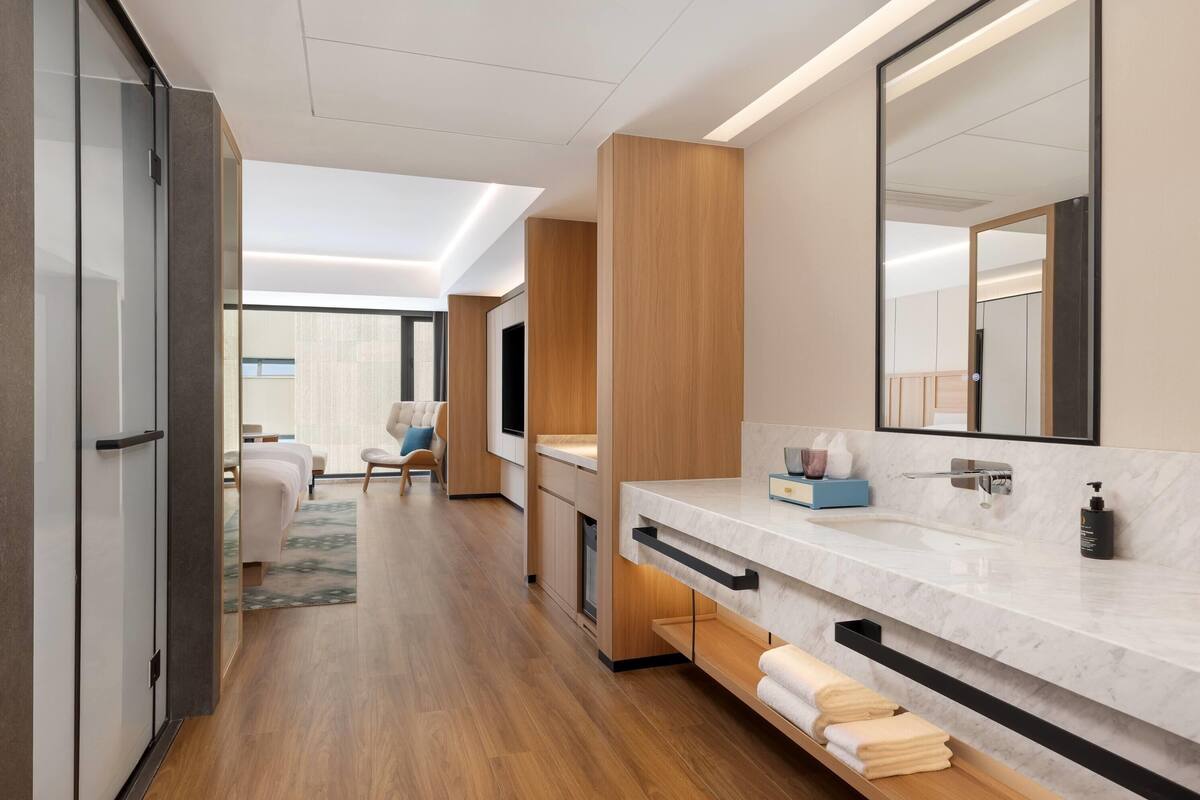 Een moderne hotelkamer met een grote spiegel, een marmeren wastafel, houten vloeren en een zithoek met een bank en stoelen.