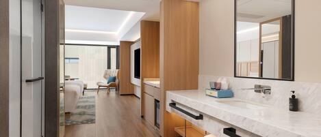 A modern hotel room with a large mirror, a marble sink, wooden flooring, and a seating area with a sofa and chairs.