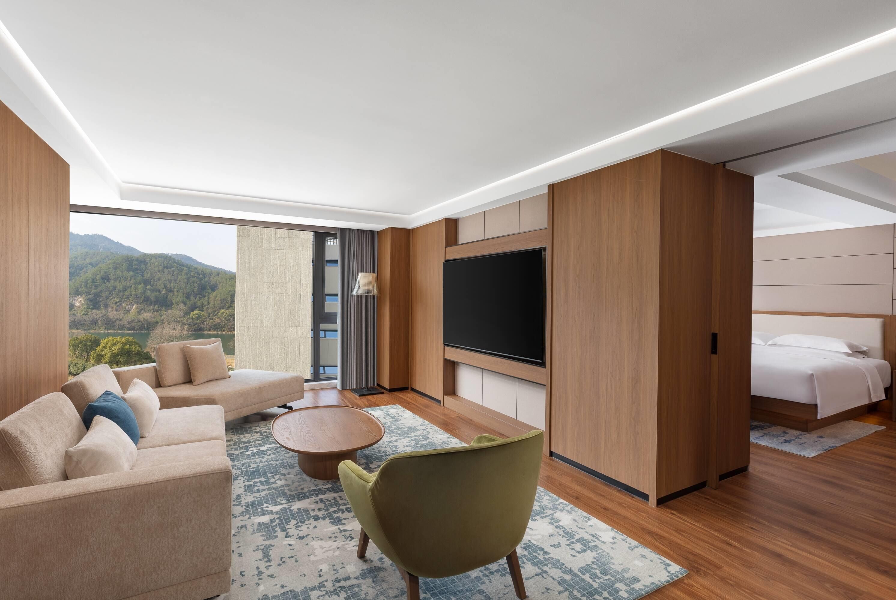 A modern hotel room with a large window, a flat-screen TV, a sofa, a chair, and a bed.