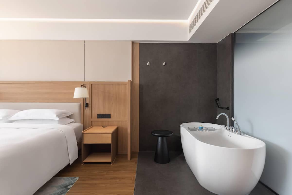 Een moderne hotelkamer met een groot bed, een nachtkastje, een klein krukje, een ruim bad en een wastafel aan de muur.