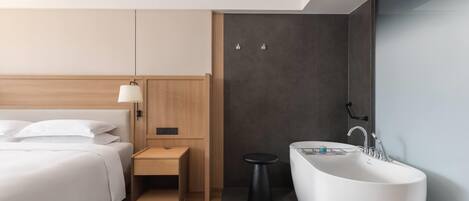 A modern hotel room with a large bed, a bedside table, a small stool, a large bathtub, and a wall-mounted sink.