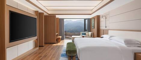 A modern hotel room with a large flat-screen TV, a bed, a seating area, and a view of mountains.