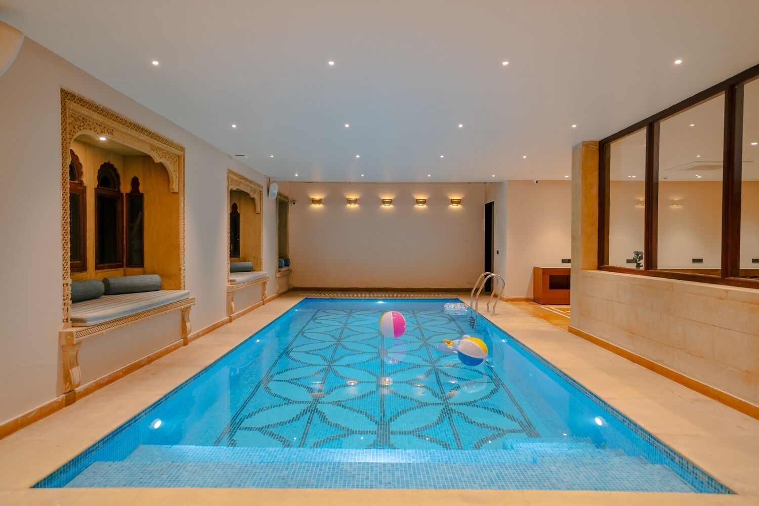 An indoor swimming pool with a geometric tile pattern, seating areas with cushions, and wall-mounted lighting.