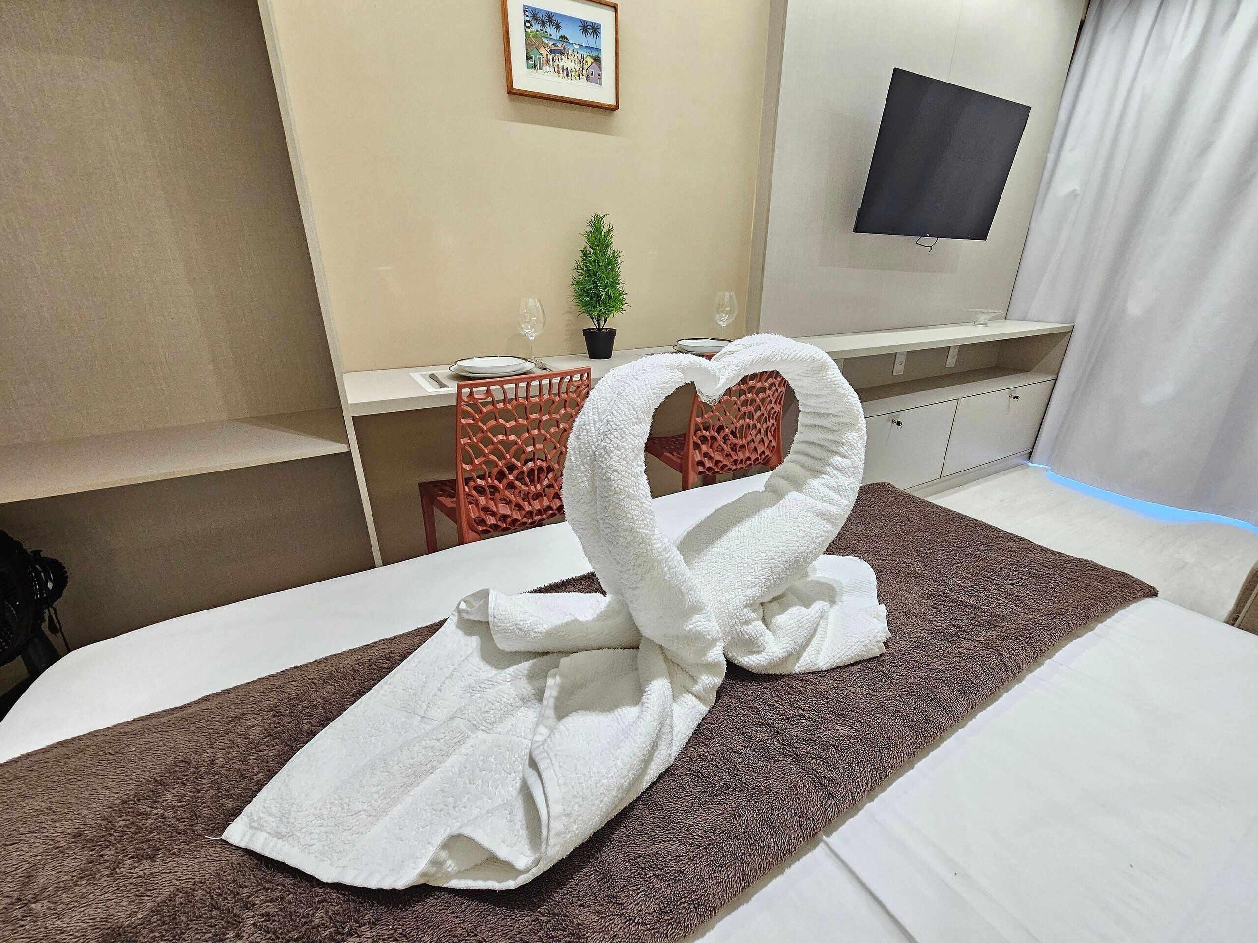 A hotel room with a neatly made bed, a desk with a chair, a small potted plant, and a TV mounted on the wall.