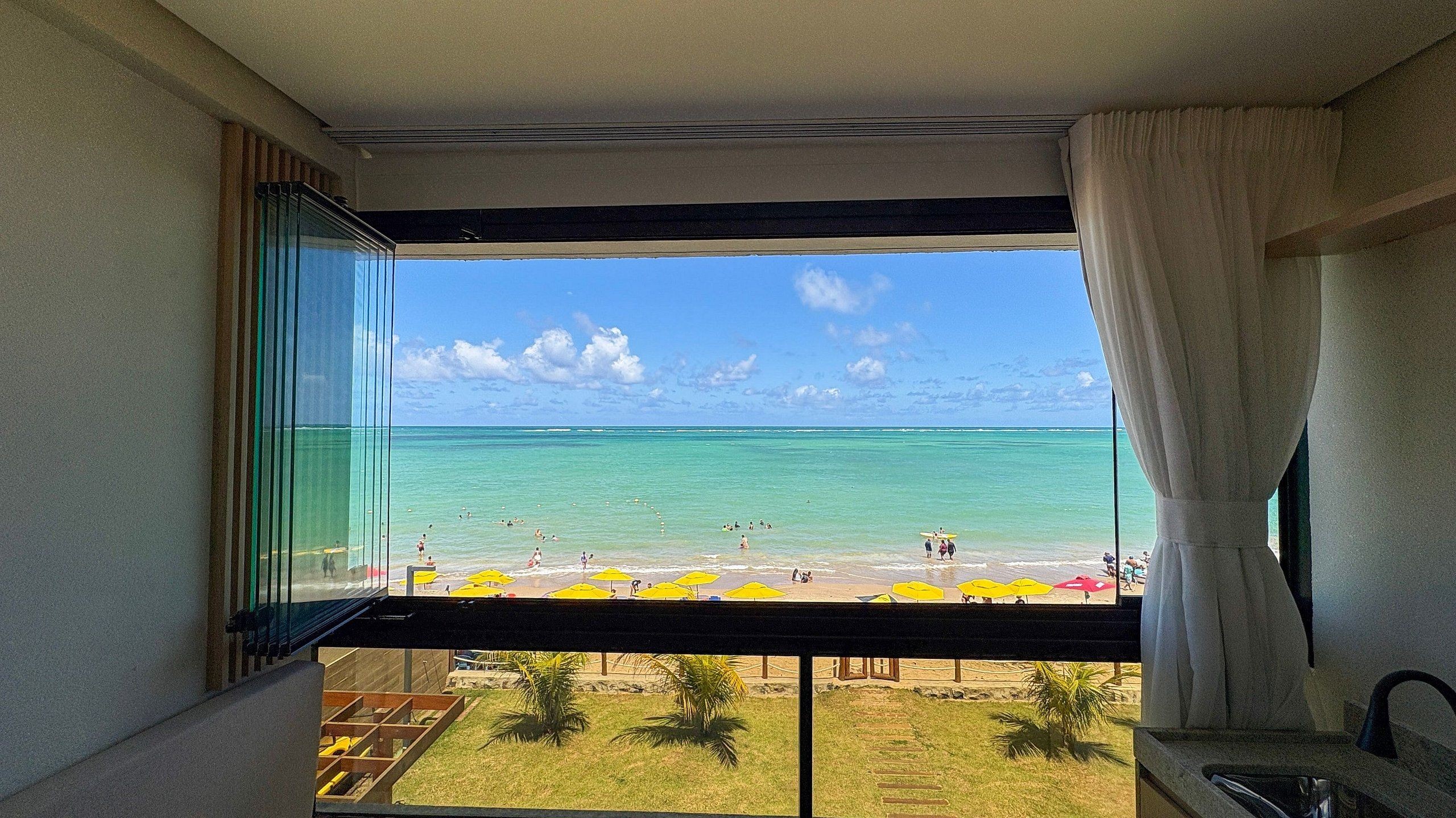 A room with a large window overlooking a beach with umbrellas and people.