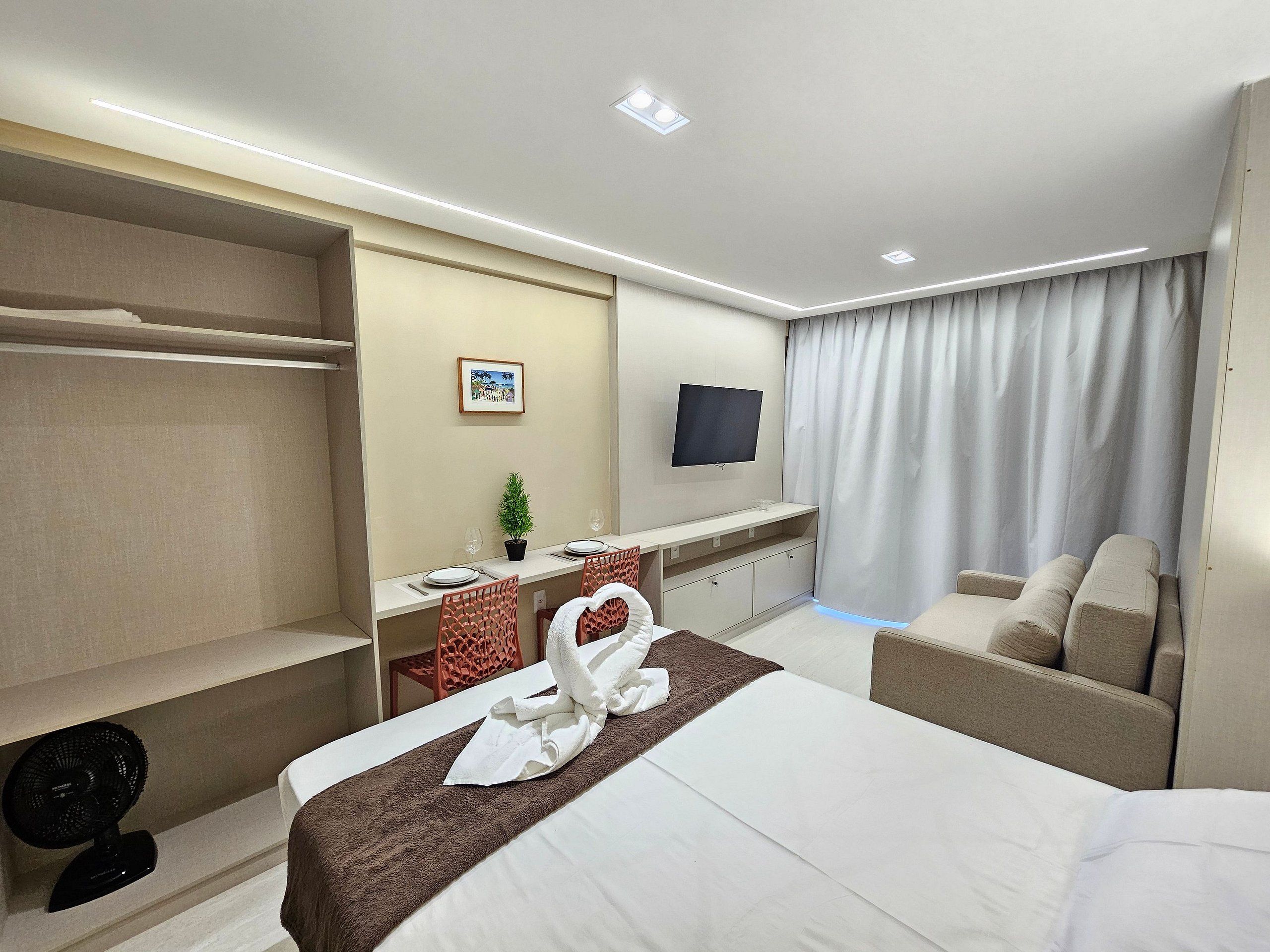 A hotel room with a bed, a desk with a flat-screen TV, a chair, a fan, and a window with curtains.