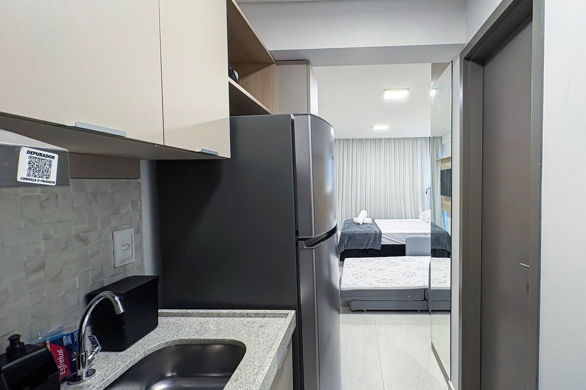 A compact hotel room with a kitchenette, a bed with a mattress and pillows, a bedside table, and a telephone.