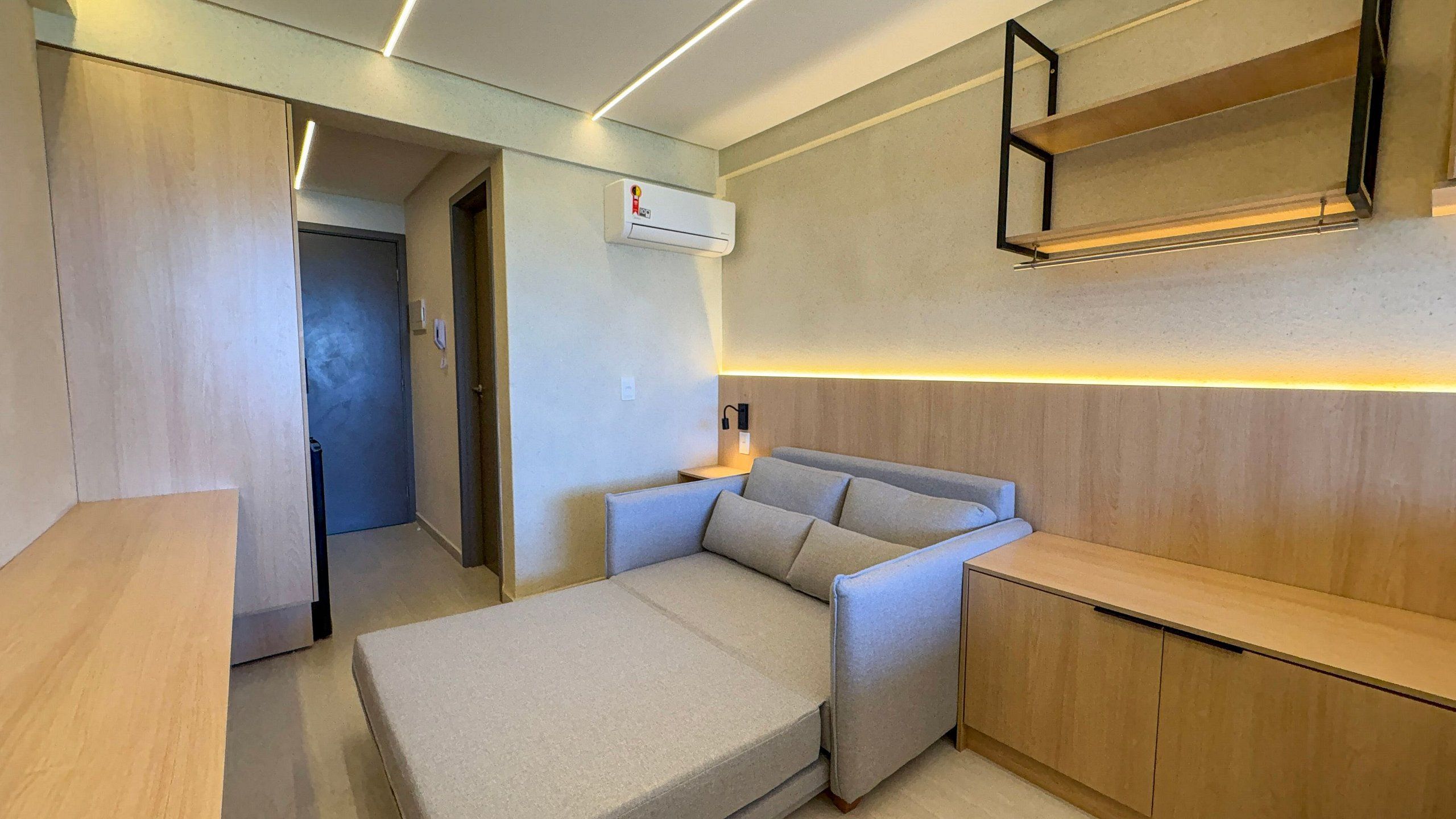 A compact room with a bed, a bedside table, a wall-mounted shelf, and a door leading to another room.