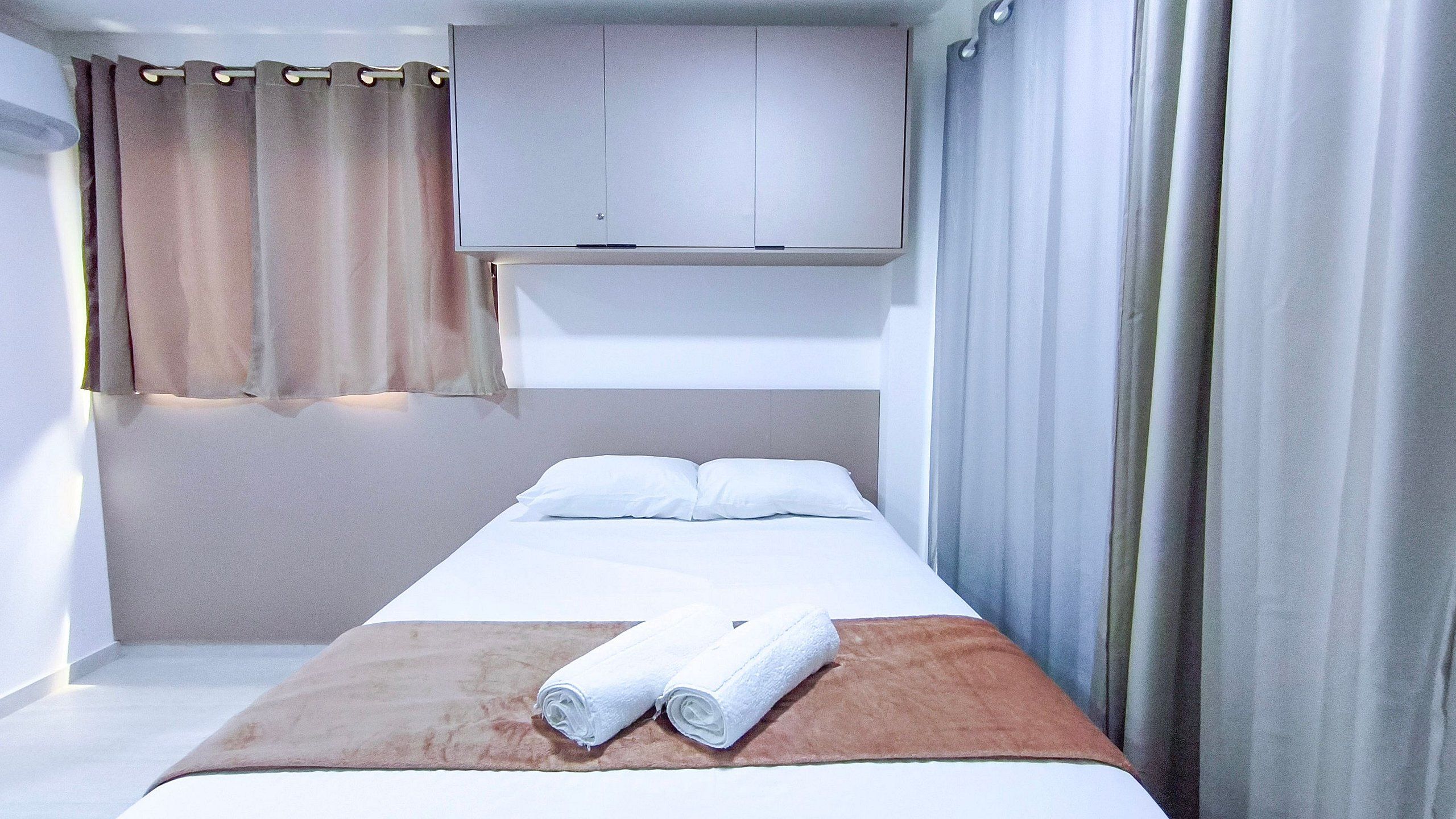 A single bed with a headboard, two pillows, a brown blanket, and two rolled towels.