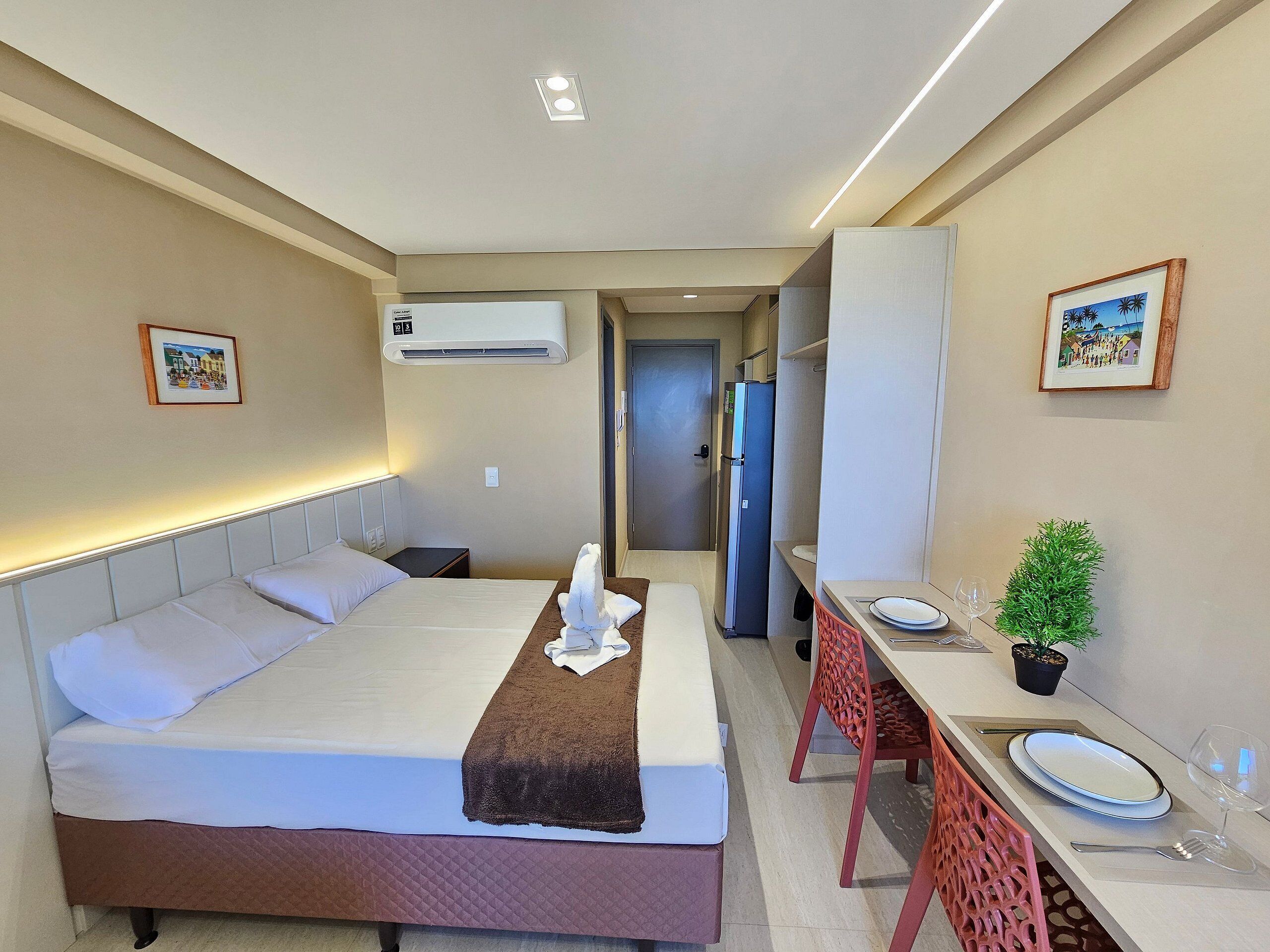 A hotel room with a bed, bedside table, air conditioning unit, framed pictures, and a dining area with a table and chairs.