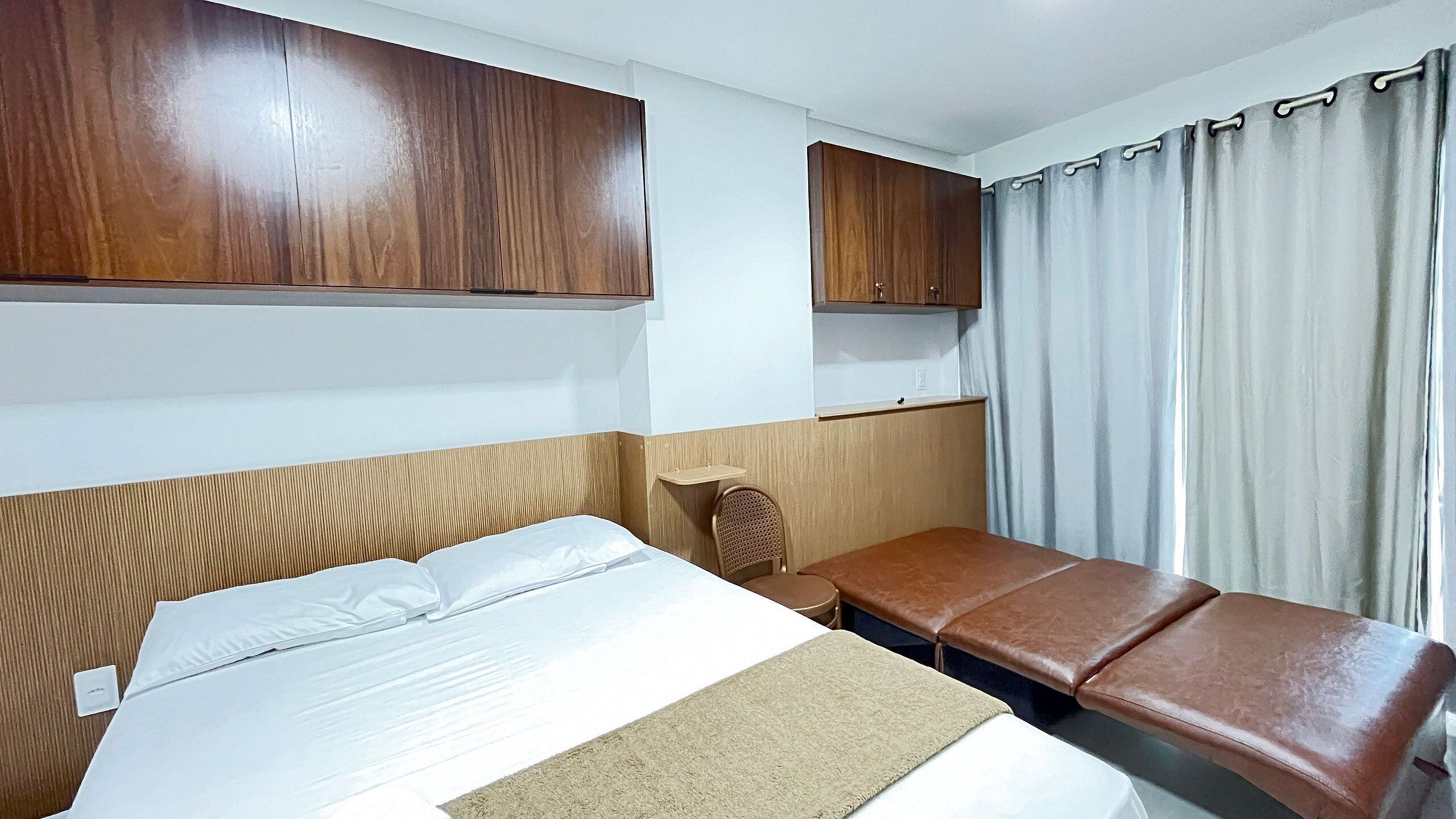 A hotel room with a bed, a small table, a chair, and a window with curtains.