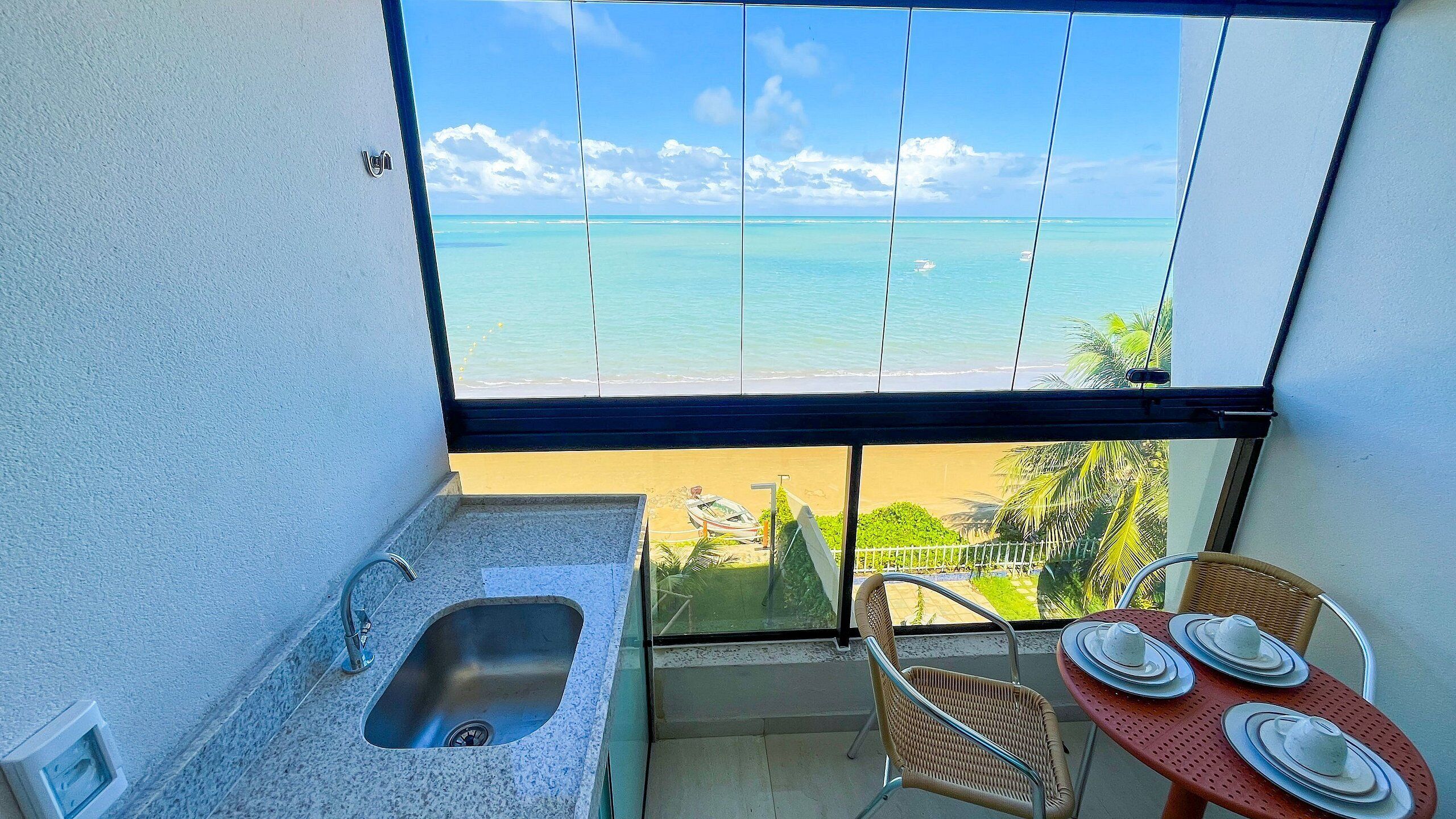 A balcony with a view of the ocean, a sink, a chair, and a table with plates and cups.