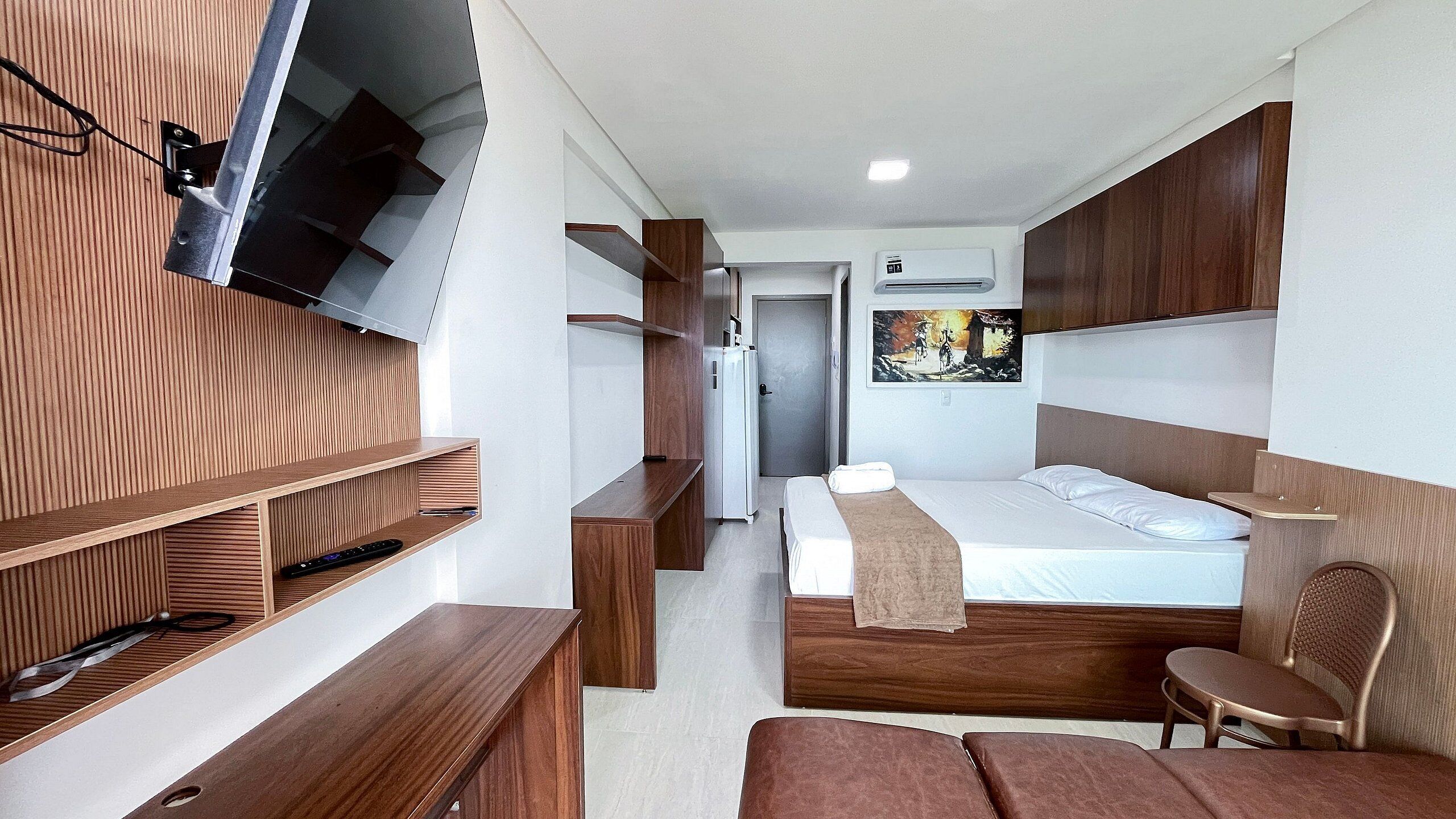 A modern hotel room with a bed, wooden furniture, a TV mounted on the wall, and a painting on the wall.