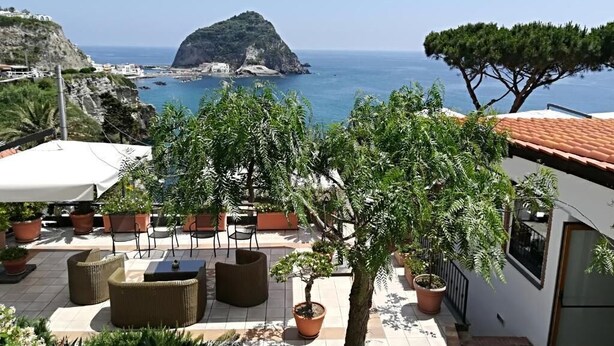 A terrace with outdoor seating, a view of the sea, and a mountainous island in the distance.