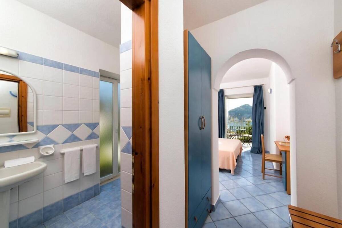 A bathroom with a sink, mirror, and towel rack, leading to a bedroom with a bed and a table.