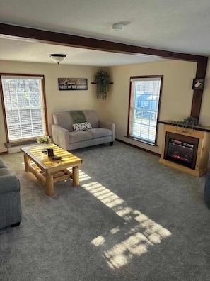 A living room with a sofa, a wooden coffee table, a fireplace, and large windows. - Cozy Cottage Sleeps 4, Walk to Downtown Springs (Berkeley Springs)