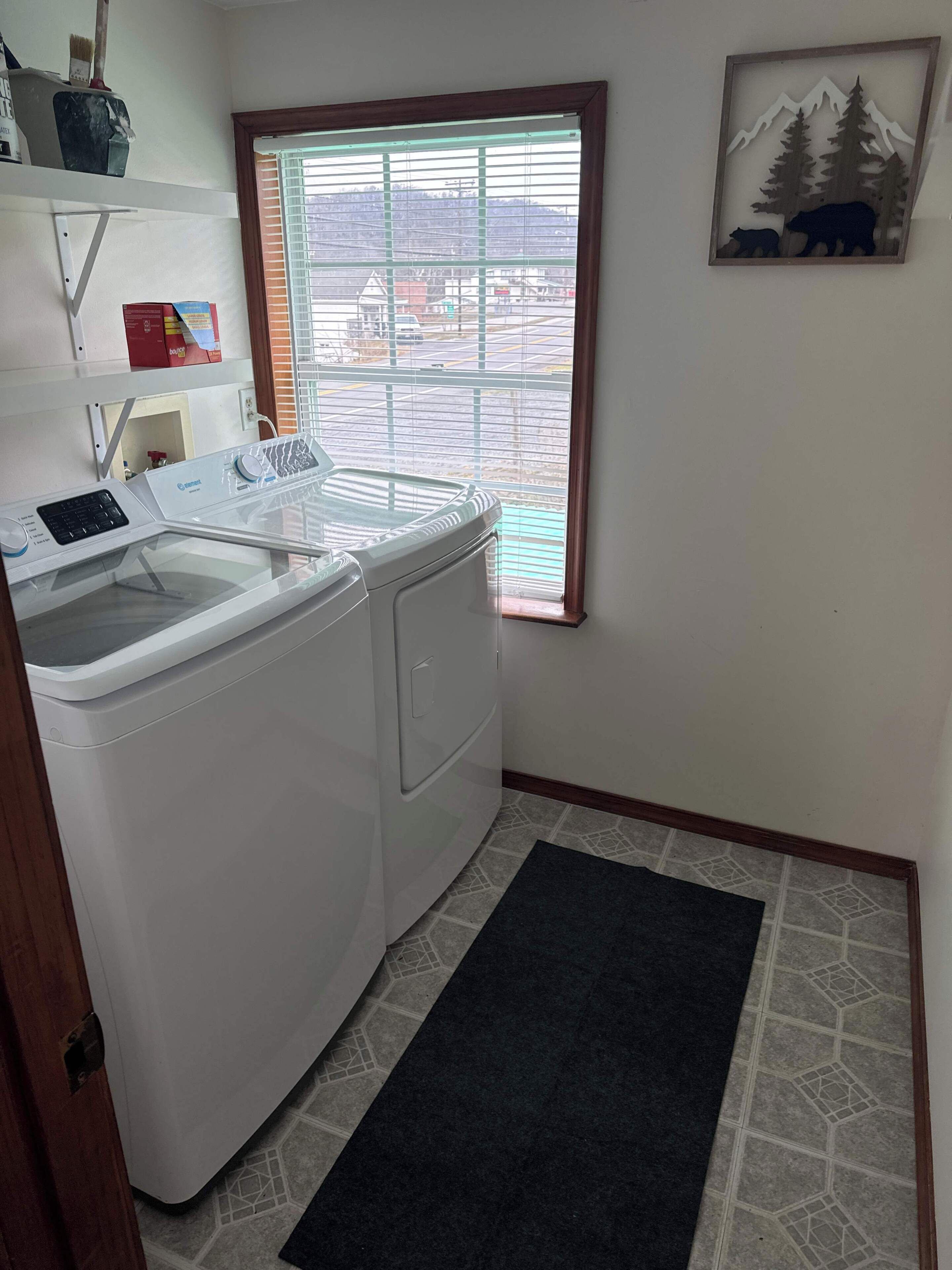 Une buanderie équipée d’un lave-linge et d’un sèche-linge à chargement frontal, d’une fenêtre avec des stores, d’une porte et d’un petit tapis sur le sol.