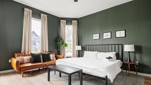 A bedroom with a bed, a sofa, a bedside table, a lamp, and a window with curtains. - Indy Getaway - Near Downtown Indy - Sleeps 10! (Indianapolis)