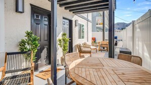 Een overdekt terras met een houten tafel en stoelen, een klein tuintje met planten, en een deur met een zwarte omlijsting.