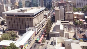 Un paisaje urbano con una torre destacada, un edificio grande de techo plano y una calle concurrida con coches y un autobús.