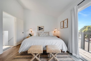 Ein Schlafzimmer mit einem großen Bett, zwei Nachttischen mit Lampen, einer gewobenen Bank und Blick ins Freie durch ein Fenster.