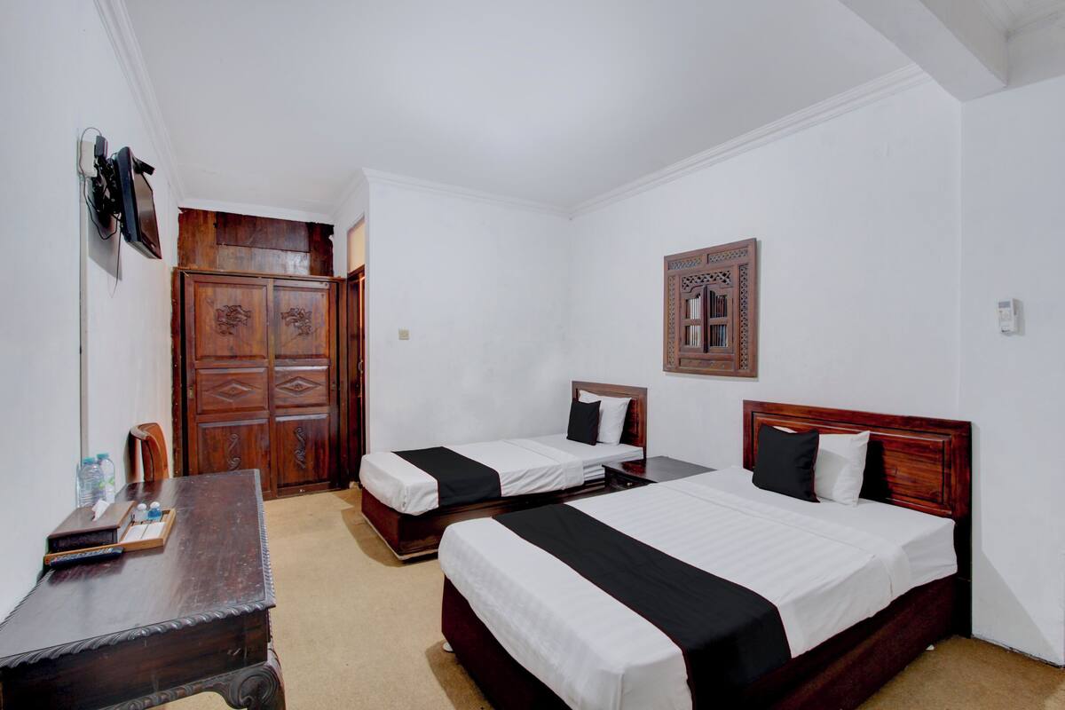 A hotel room with two single beds, a wooden wardrobe, a desk with a chair, and a small table with a bottle and glasses.