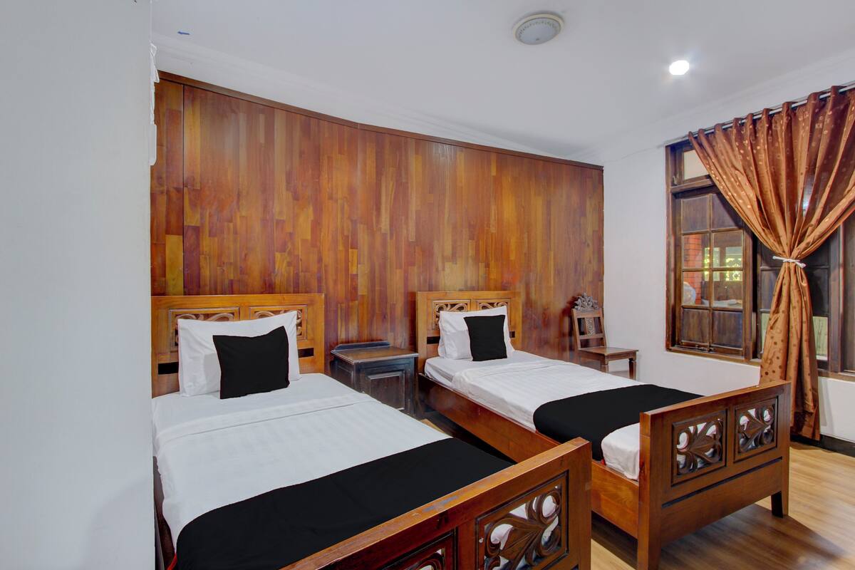 A hotel room with two wooden beds, a wooden headboard, a nightstand, and a window with curtains.